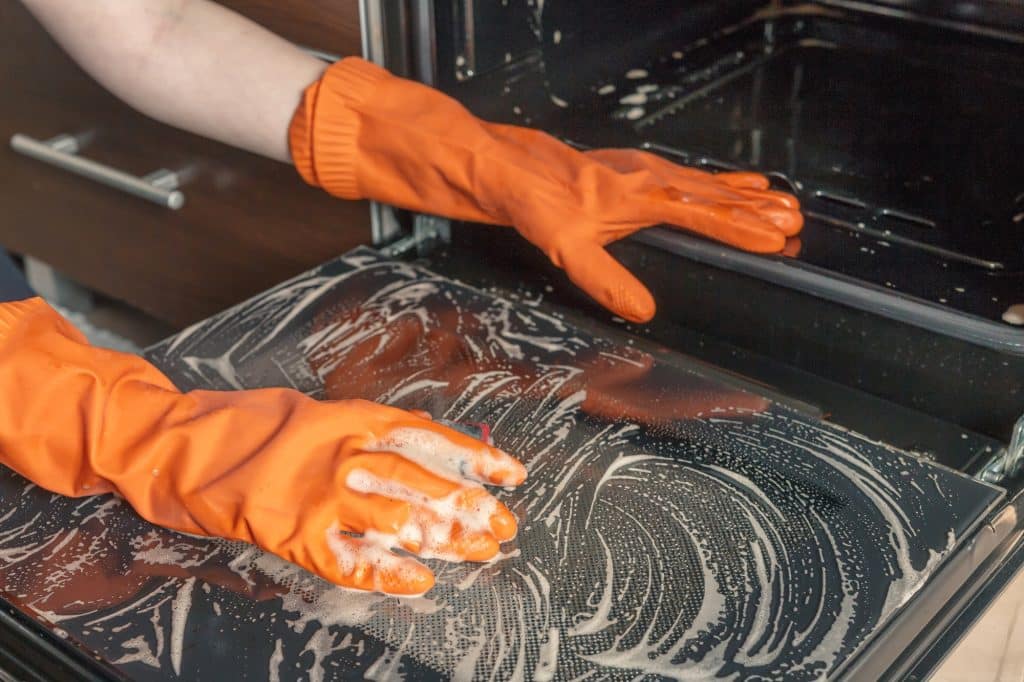Handen in handschoenen die oven schrobben met schoonmaakmiddelen | Hoe keuken schoonmaken | Asenso Schoonmaakdiensten