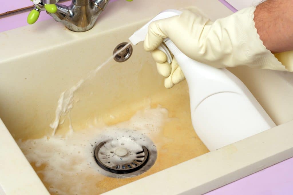 Hand met rubberen handschoen spuit reinigingsoplossing op een vuile gootsteen | Hoe roestvlekken badkamer schoonmaken | Asenso Schoonmaakdiensten