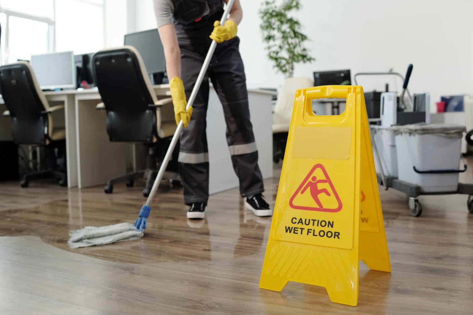 Geel waarschuwingsbord op gedweilde vloer terwijl iemand op de achtergrond dweilt | Vloerbedekking schoonmaken kantoor | Asenso Schoonmaakdiensten