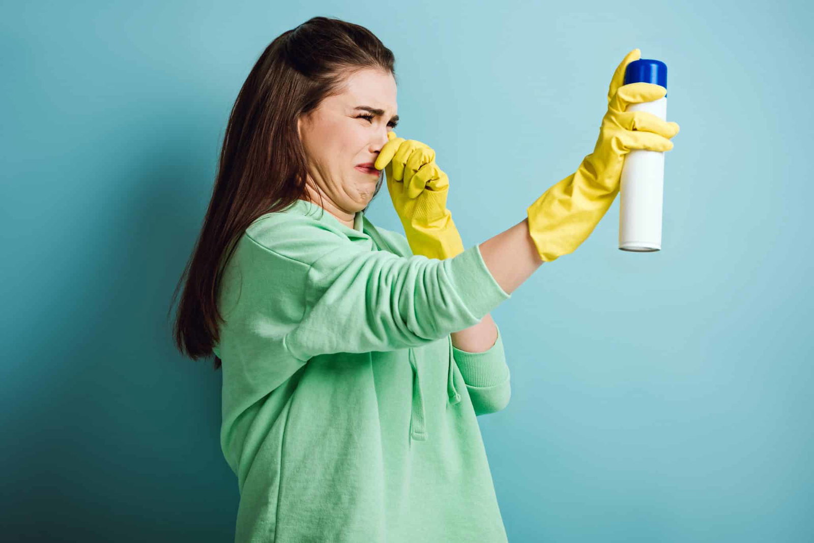Vrouw met bruin haar en gele schoonmaakhandschoenen die haar neus dichtknijpt en een spray spray, ze kijkt vies | Geur verwijderen uit huis | Asenso Schoonmaakdiensten