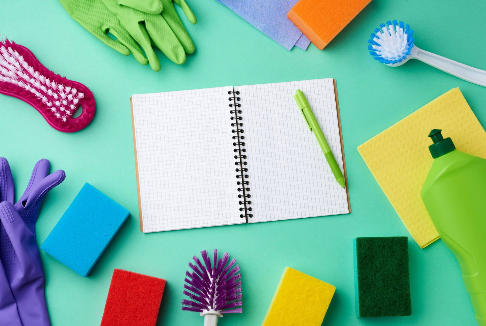 Close-up van een divers aantal schoonmaakartikelen zoals zeep, spons, handschoenen, schrobber, met in het midden een opengeslagen, leeg notitieboek met een pen erop | Schoonmaakschema maken | Asenso Schoonmaakdiensten