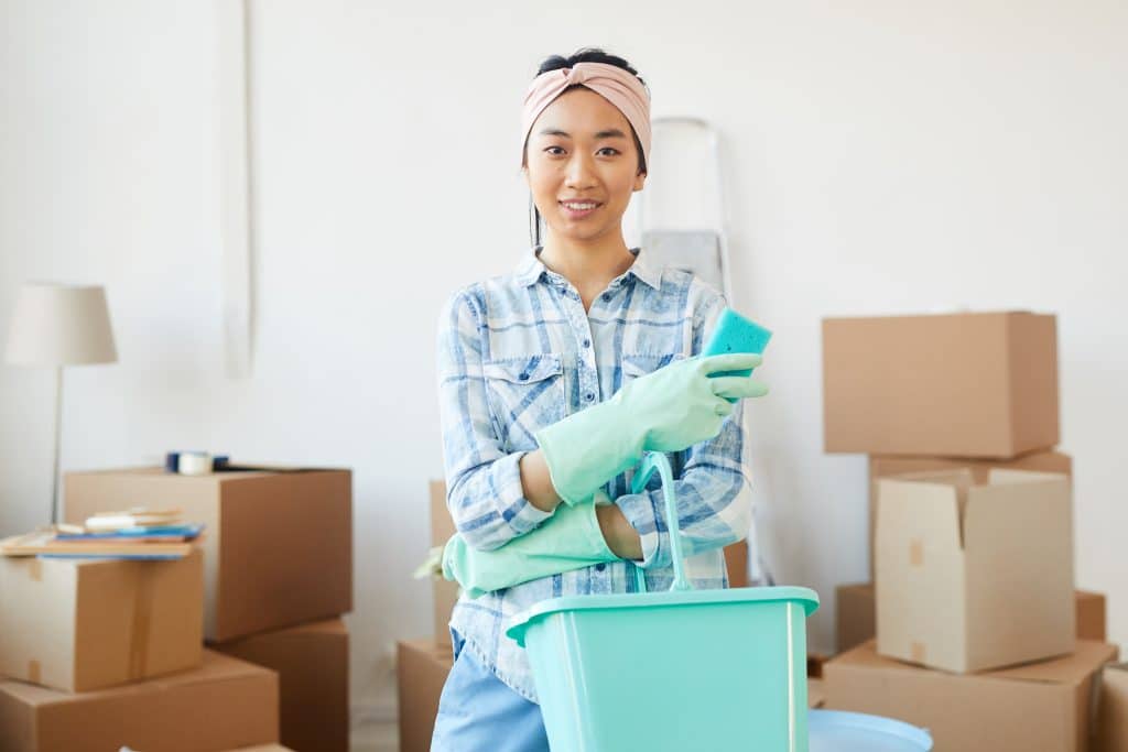 Vrouw met schoonmaakmiddelen die lachend te midden van verhuisdozen staat | Huis schoonmaken voor verkoop | Asenso Schoonmaakdiensten