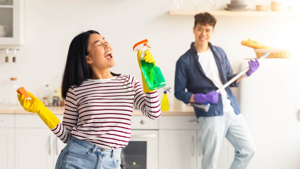 Vrolijk koppel dat de keuken schoonmaakt terwijl de vrouw doet alsof ze in de spray zingt, terwijl de man een dweil als gitaar vasthoudt | Voorjaarsschoonmaak checklist | Asenso Schoonmaakdiensten