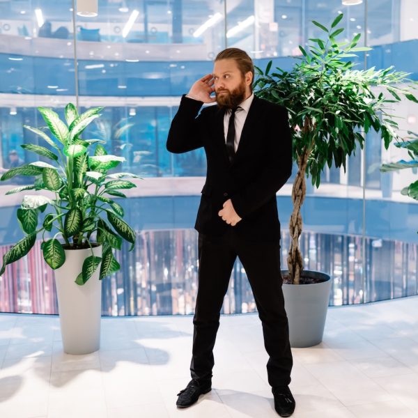 Full body of serious adult male security guard wearing formal suit listening to message in earpiece while standing in modern business center with glass walls